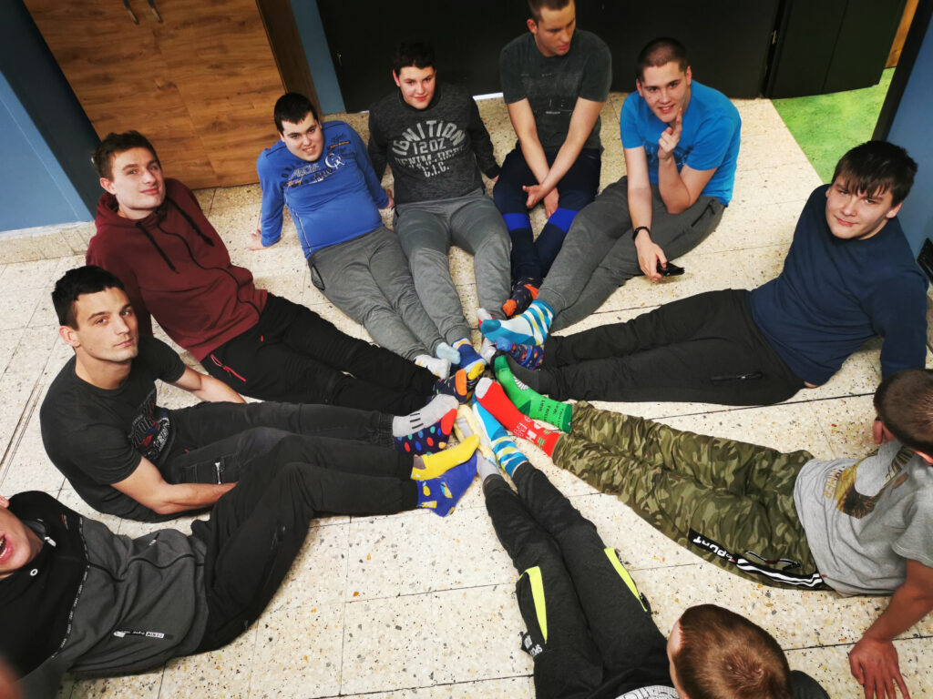 Grupa chłopaków siedzi na podłodze w kręgu. Stykają się stopami, na których mają różnokoolorowe skarpetki. 