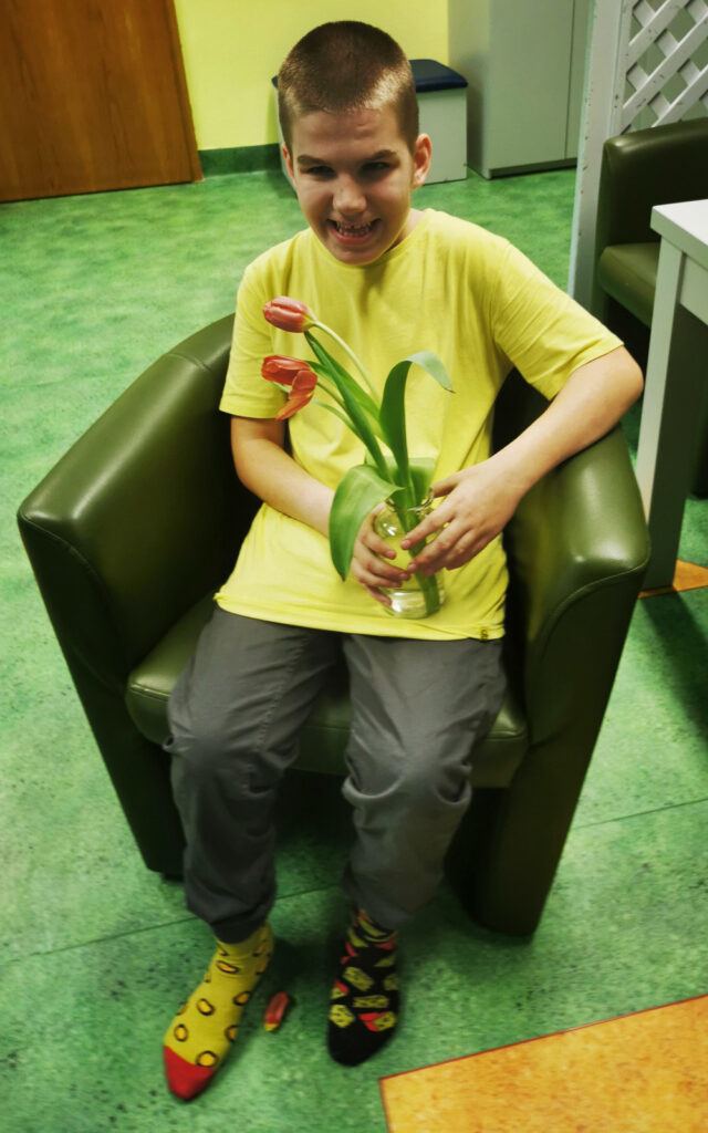 Chłopiec siedzi w fotelu, w rękach trzyma wazon z kwiatami. Szeroko się uśmiecha. Na stopach ma dwie różne skarpetki. 