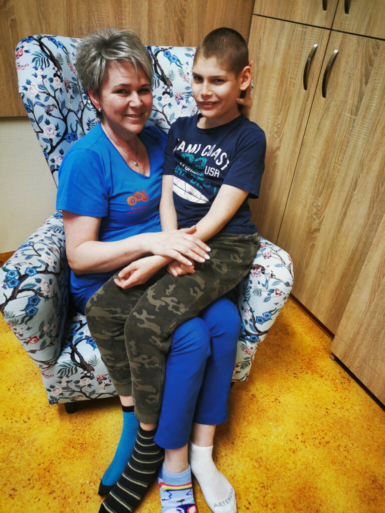 Kobieta siedzi w fotelu, a na kolanach trzyma chłopca. Oboje mają różne kolorowe skarpetki na stopach. 