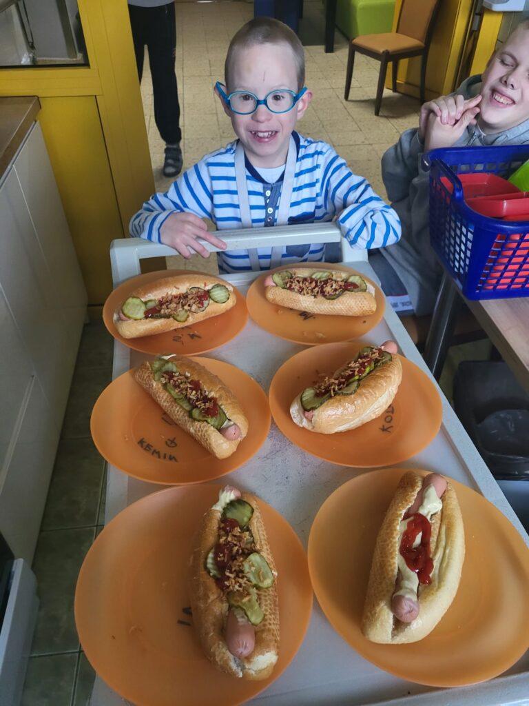 Mały chłopiec pcha przed sobą wózek, na którym ułożone są talerze z hot dogami. 