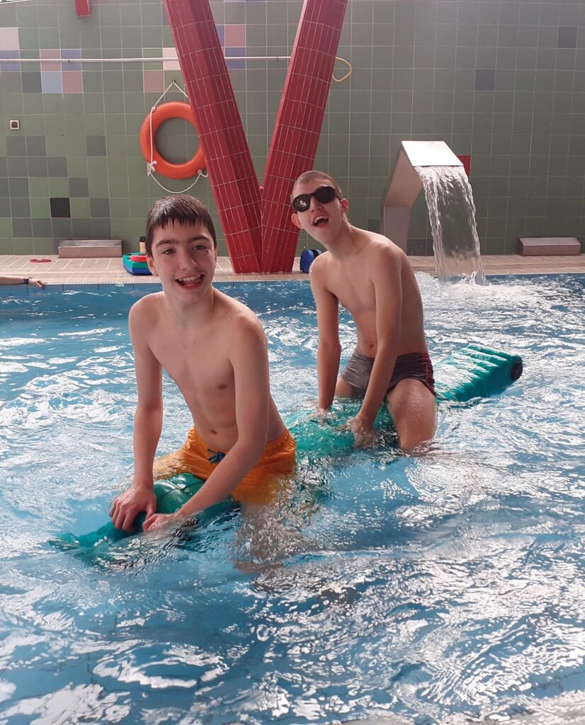 dwóch uśmiechniętych  chłopaków w basenie siedzących na pływającej po 
 wodzenie konstrukcji. 