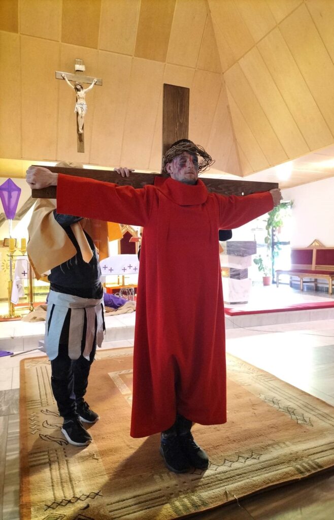 Mężczyzna w czerwonym stroju i koronie cierniowej na głowie stoi z rozłożonymi na boki rękami. Plecami przylega do stojącego za nim krzyża. 