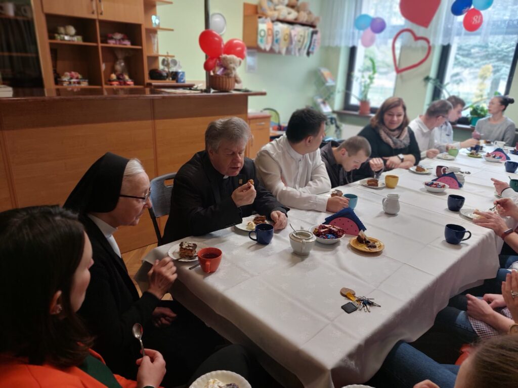 Grupa osób siedząca przy zastawionym ciastami, cukierkami i kawą stole. 