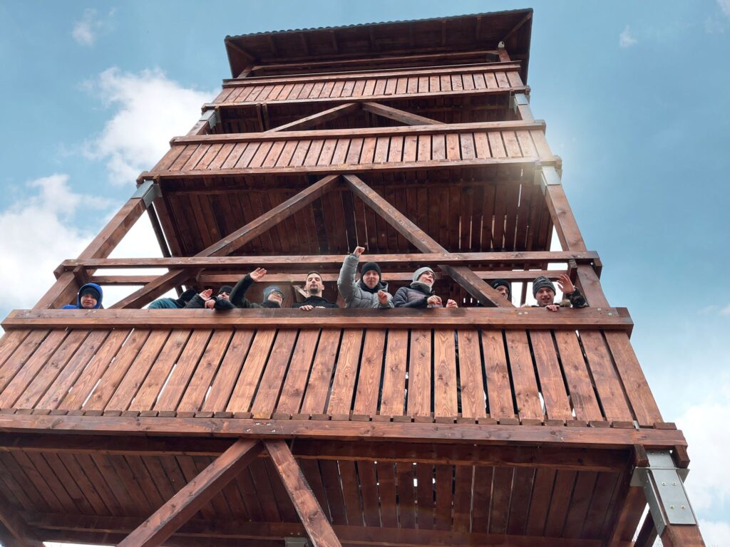 Grupa osób wychyla się z drewnianej wieży i macha do fotografa. 