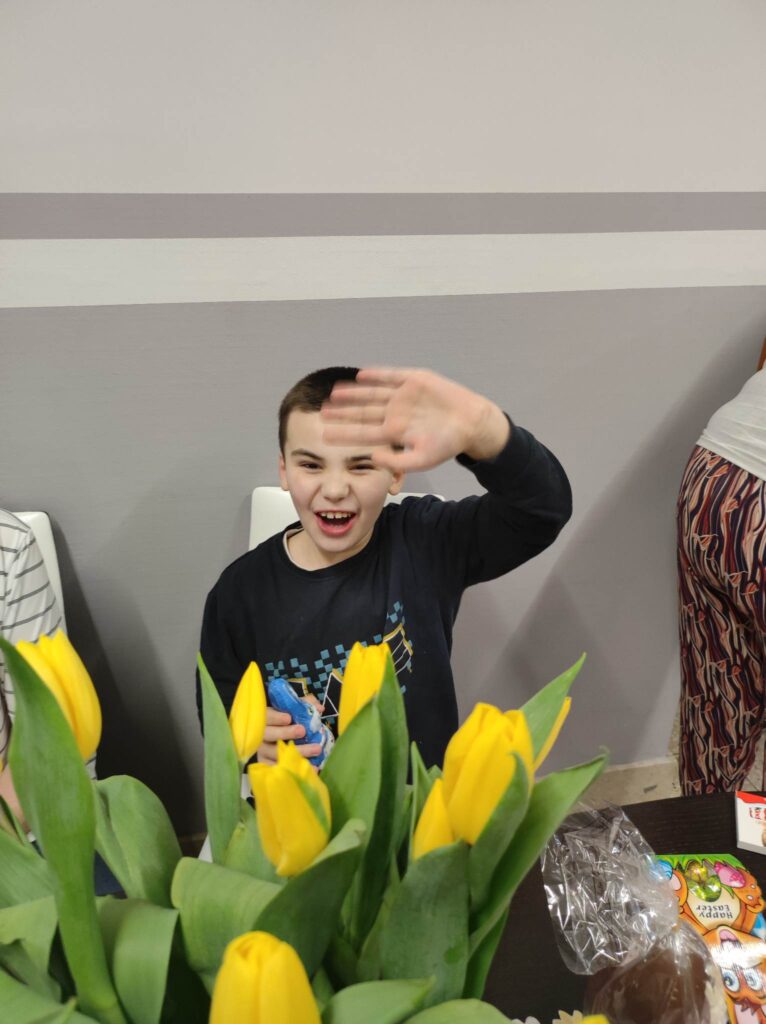 Chłopiec macha. Przed nim wazon z żółtymi tulipanami. 