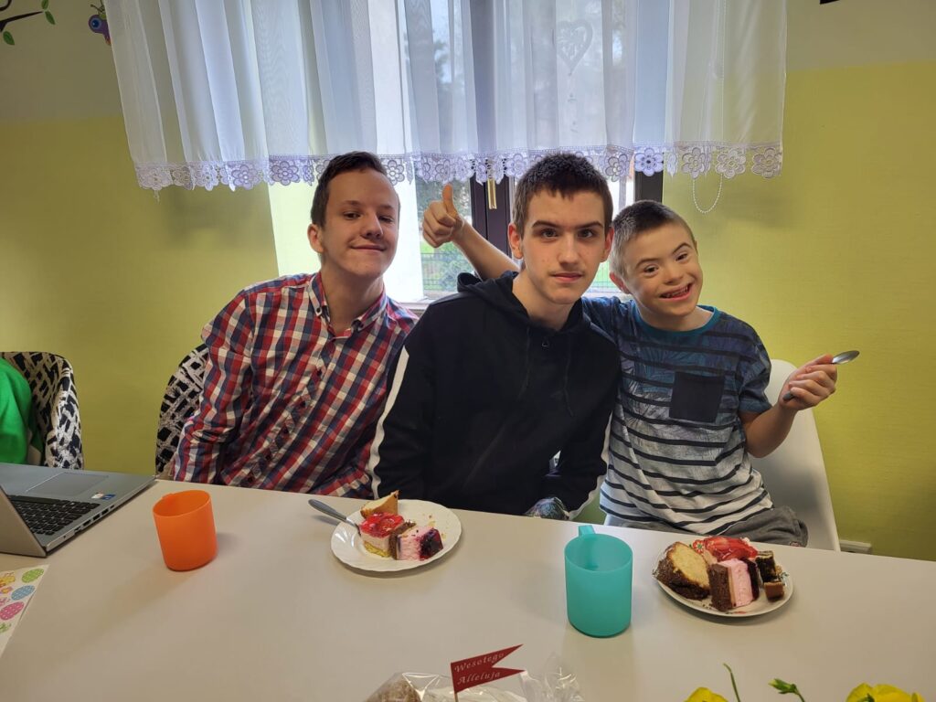 Trzech chłopaków siedzących za stołem. Przed nimi talerzyki z ciastami. 
