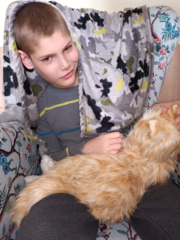 Owinięty w koc chłopiec trzyma kota na kolanach. 