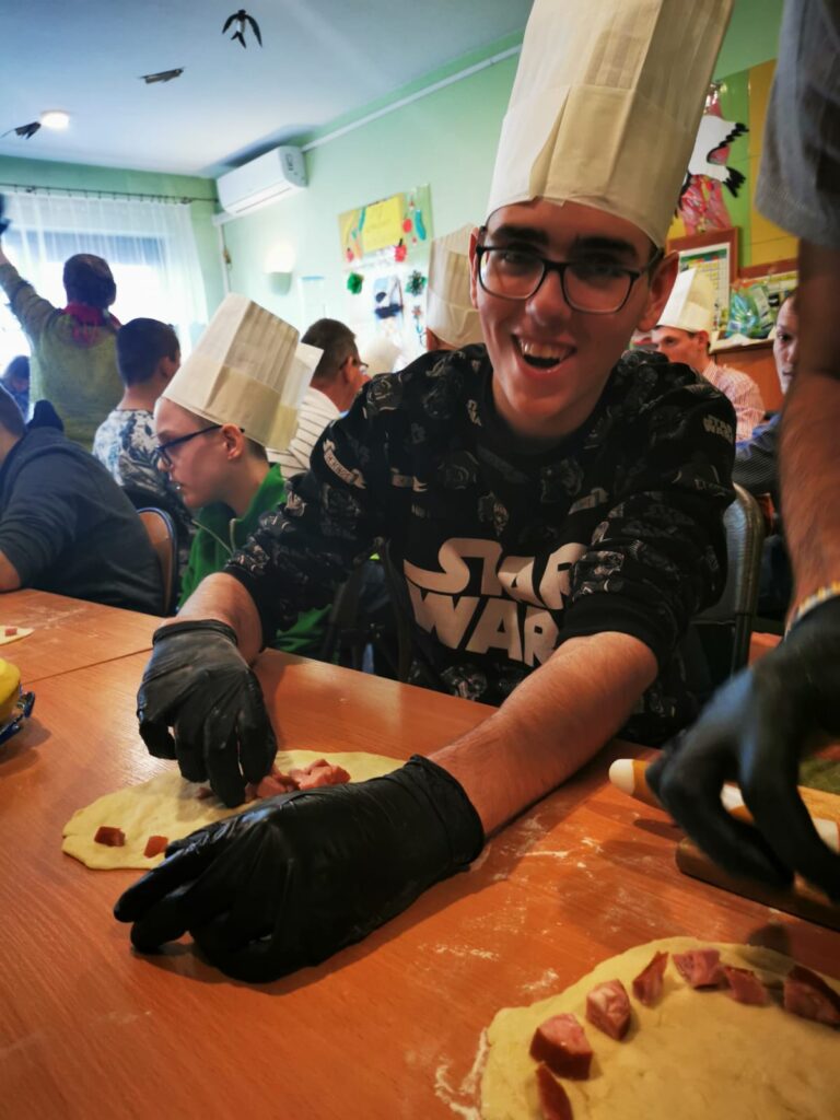 Uśmiechnięty chłopak w czapce kucharskiej na głowie układa na cieście kawałki kiełbasy i boczku. 