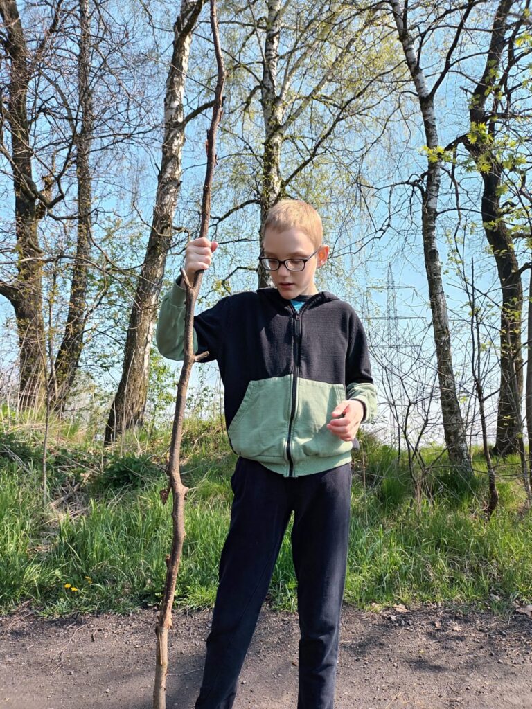 Chłopak stojący na tle drzew. W ręce trzyma długi kij, którym podpiera się o ścieżkę. 