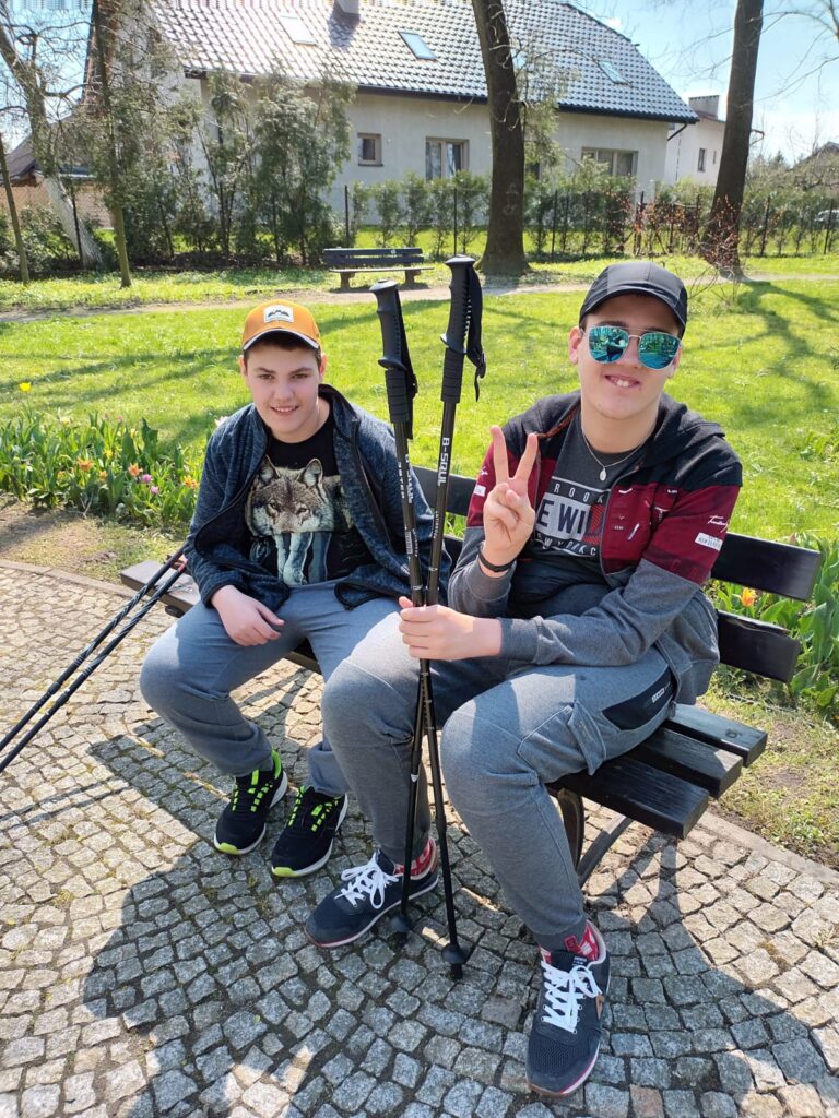 Dwóch chłopaków siedzi na ławce w parku. W rękach trzymają kijki. 
