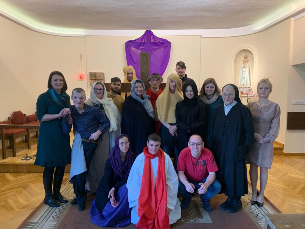 Zdjęcie grupowe w kaplicy. Ludzie w strojach teatralnych. W tle zakryty fioletowym materiałem, duży krzyż. 