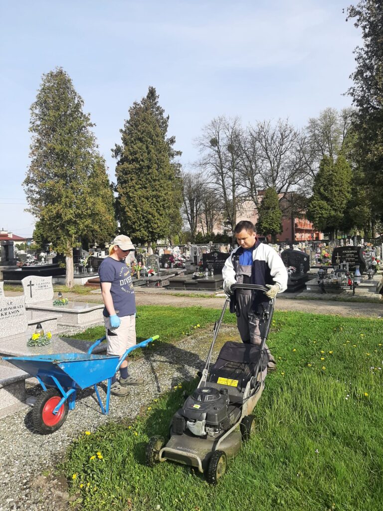 Dwóch chłopaków wykonujących pracę na cmentarzu. Jeden z nich stoi obok taczek, a drugi prowadzi kosiarkę.  