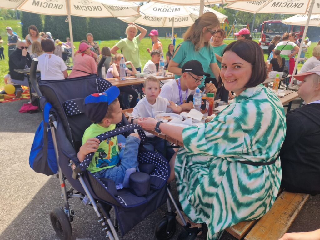 Grupa osób przy stołach pod parasolami. Kobieta podaje chłopcu siedzącemu w wózku jedzenie. 