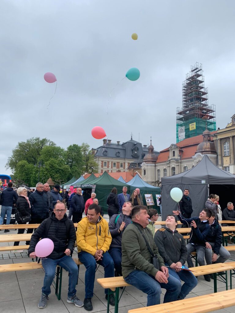 Grupa osób na ławkach pod sceną na Pszczyńskim Rynku. W tle widać fragment Pałacu. Nad głowami siedzących latają kolorowe balony.