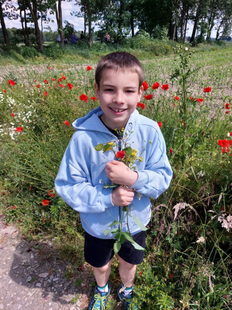 chłopiec stojący na ukwieconej łącę. Wręce trzyma bukiecik polnych kwiatów. 