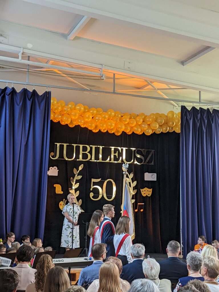 kobieta przemawiająca do mikrofonu na scenie. Za nią złoty napis "Jubileusz 50". Pod sceną ludzie siedzący na widowni. 