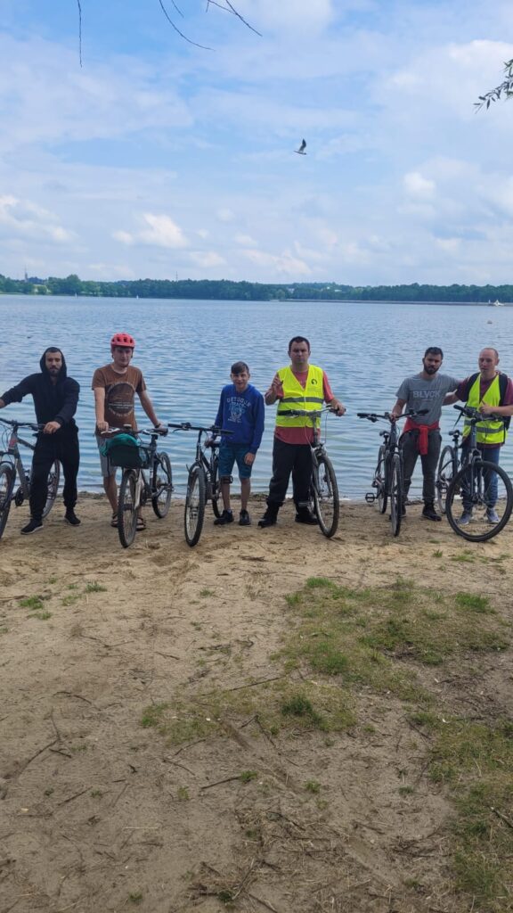 Grupa osób z rowerami pozująca do zdjęcia na brzegu jeziora. 