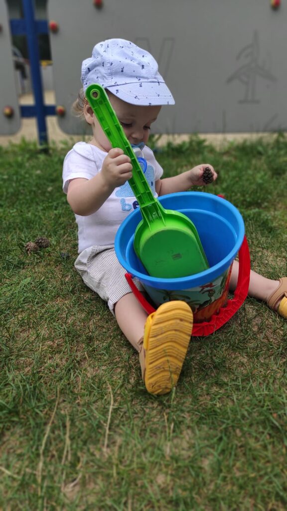 Mały chłopiec siedzący na trawie i bawiący się wiaderkiem i łopatką 