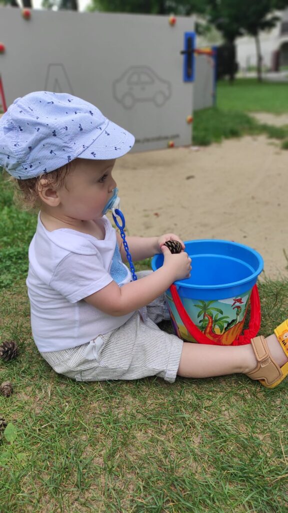 Mały chłopiec siedzący z wiaderkiem przy piaskownicy. W rękach trzyma szyszkę. 
