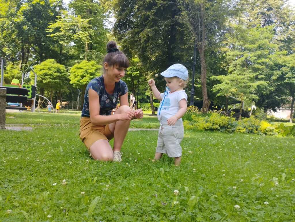 Mały chłopiec stoi na trawie, a obok kuca kobieta. Dziecko podaje jej kwiatka. 