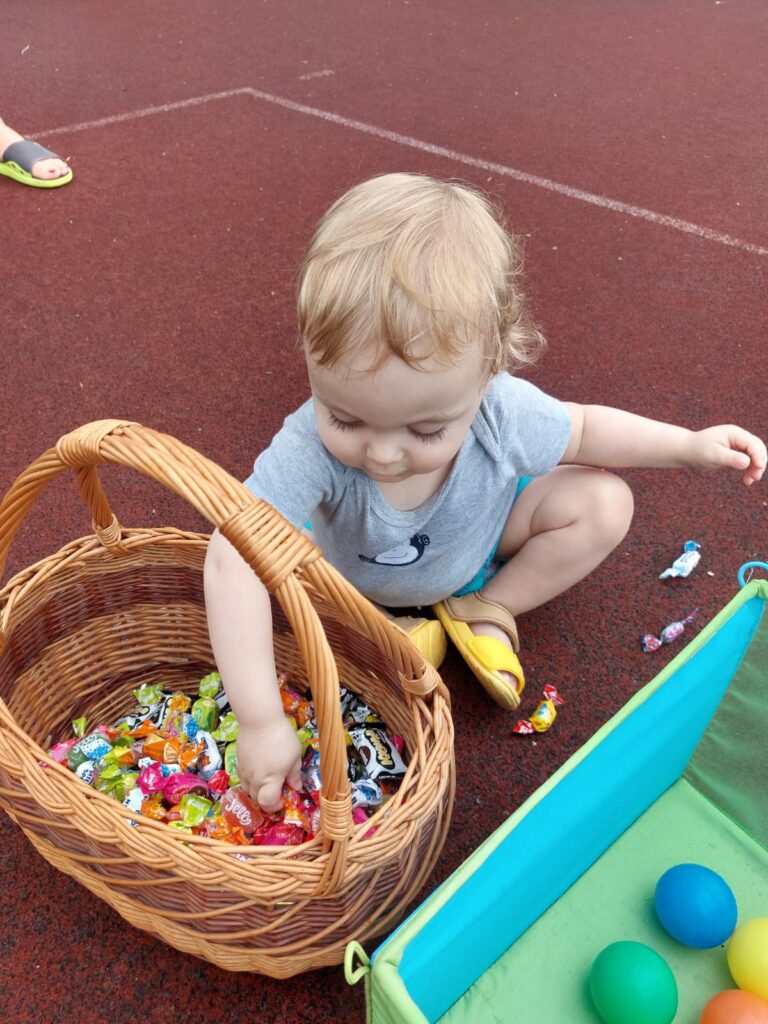 Mały chłopczyk siedzi na boisku i wyciąga cukierki z wiklinowego koszyka