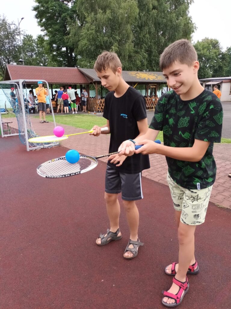 Dwaj chłopcy idący po boisku z piłeczkami niesionymi na paletkach do badmintona. Na ich twarzach wyraźnie zauważalne skupienie. 