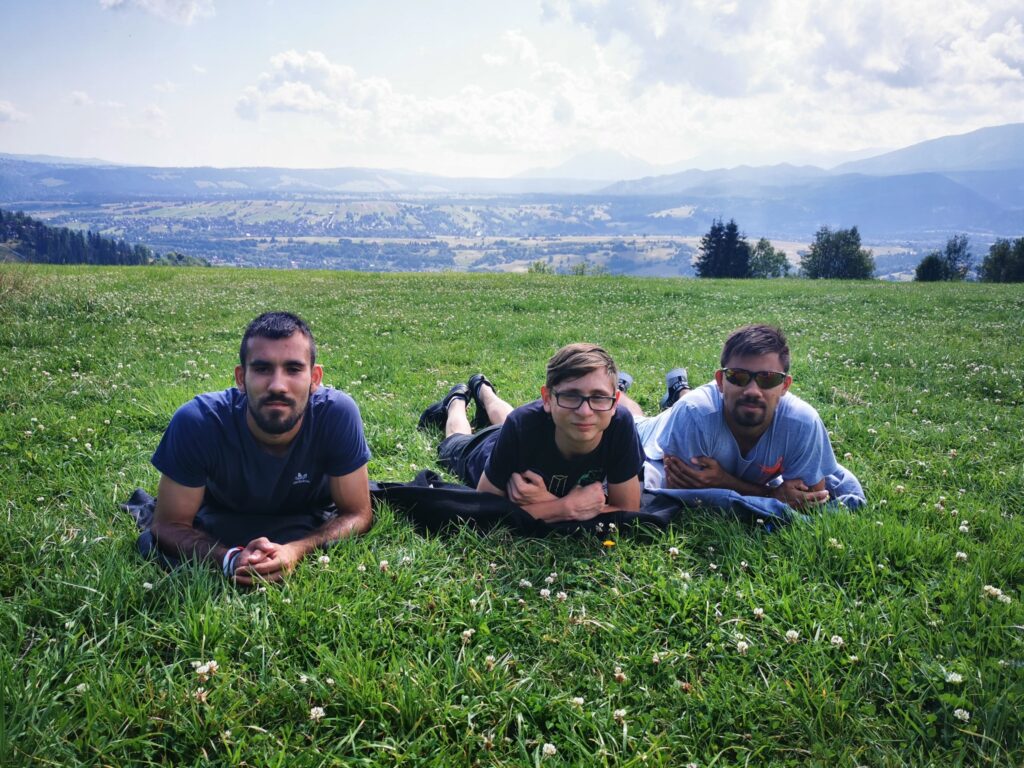 Trzech chłopaków leżących na brzuchach na trawie i spoglądających w obiektyw. Za nimi widok na łąkę i góry 