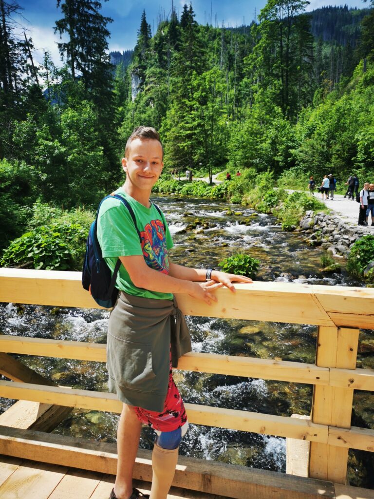 Chłopak pozujący na mostku nad górską rzeką. W tle las i góry 