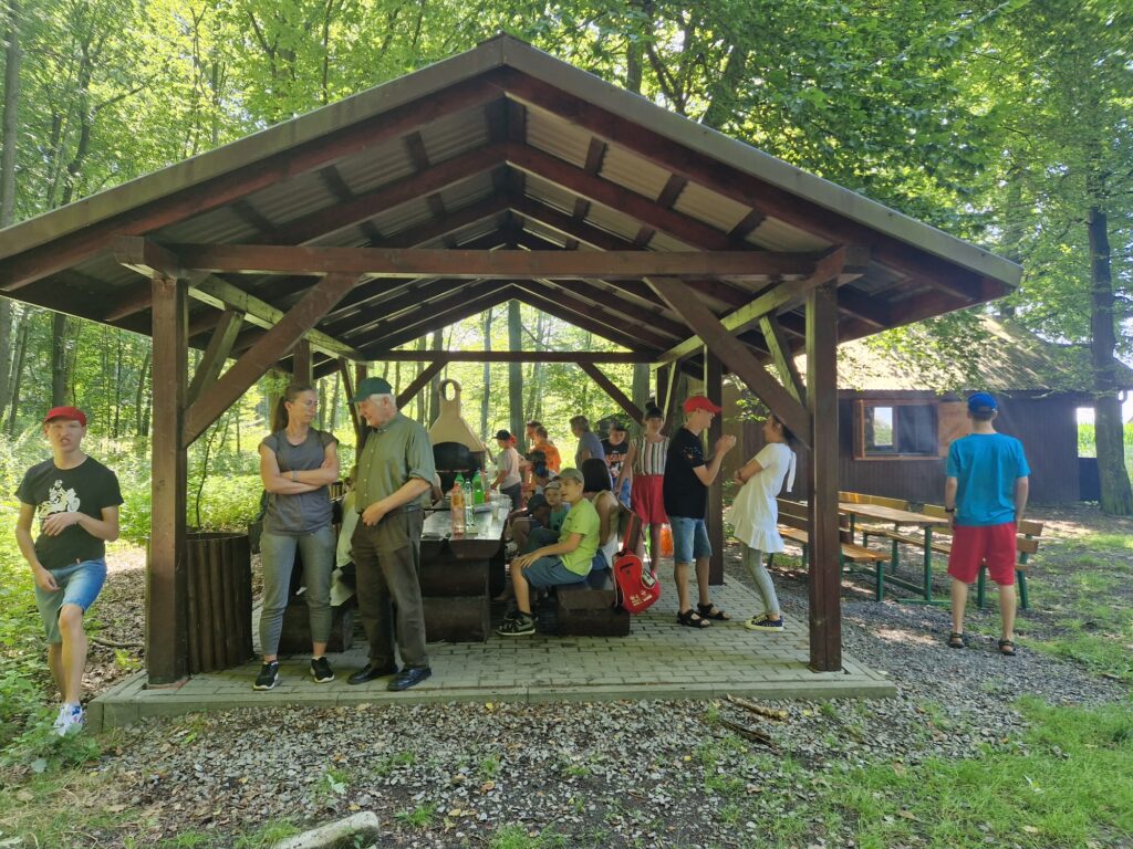 Grupa osób przy stołach pod wiatką w lesie. 