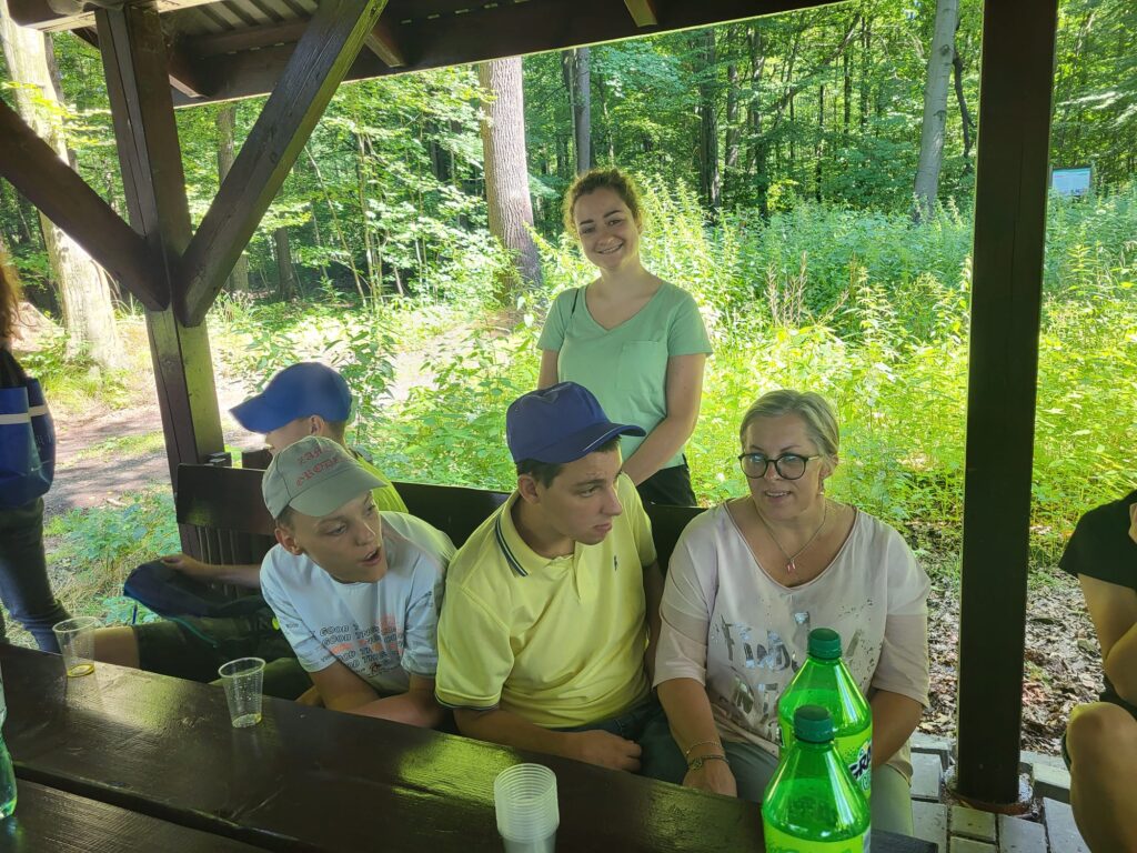 Grupa osób przy stołach pod wiatką w lesie. 