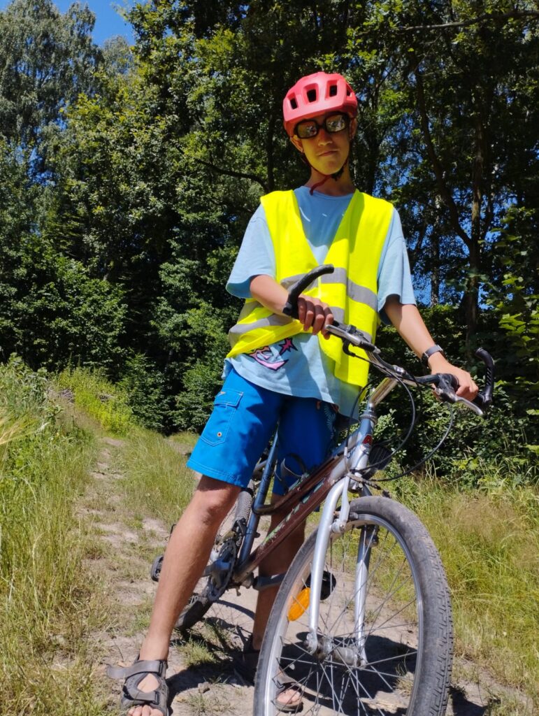 Uśmiechnięty chłopak w kasku rowerowym na głowie i w odblaskowej kamizelce stoi trzymając rower. 
