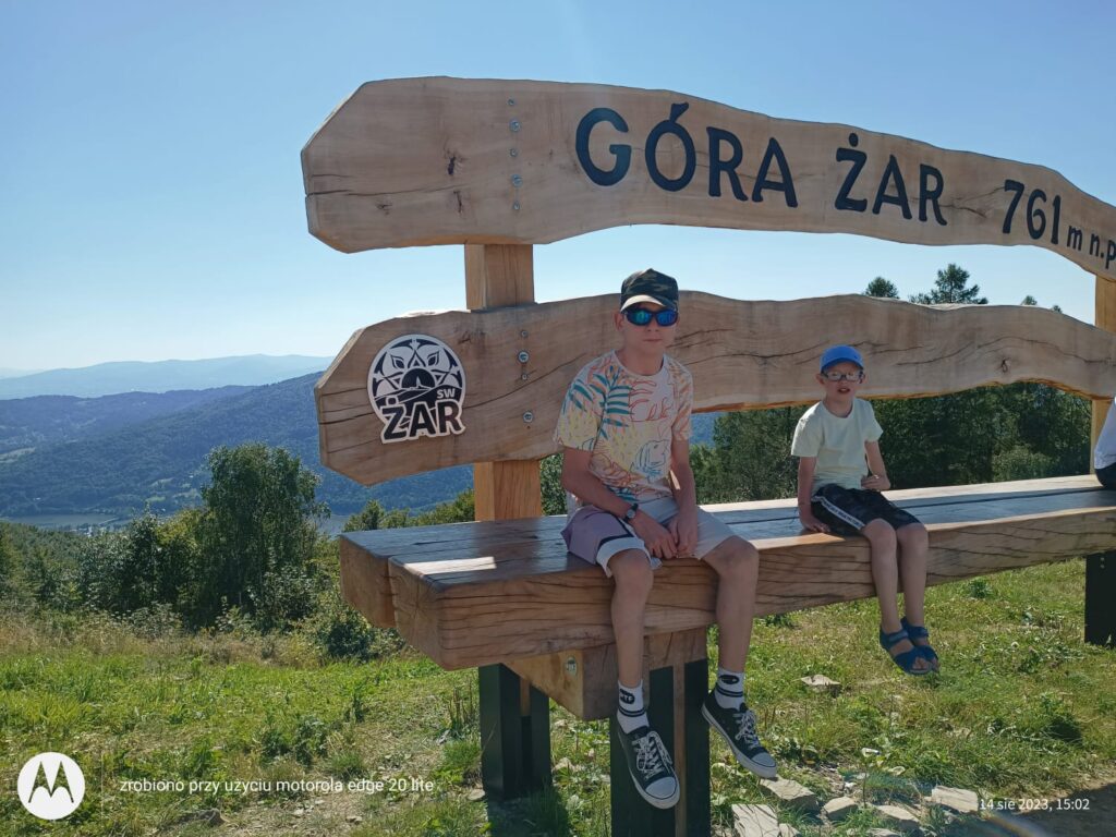 Dwóch chłopców na wysokiej, drewnianej ławce z napisem "Góra Żar" 