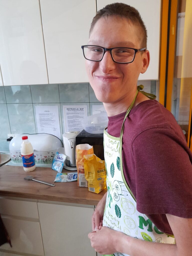 uśmiechnięty chłopiec ubrany w fartuszek stoi w kuchni. Za nim na blacie leży mąka i mleko. 