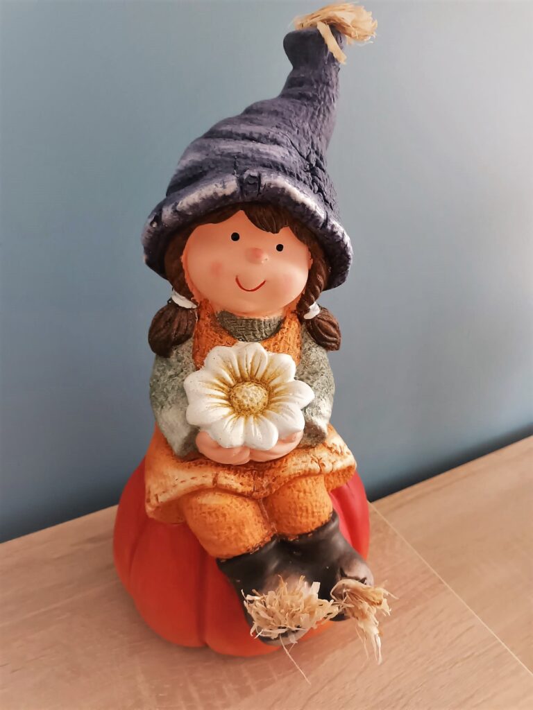 Figurka dziewczynki w sweterku i wełnianej czapce. Dziewczynka trzyma kwiat, a siedzi na dyni 