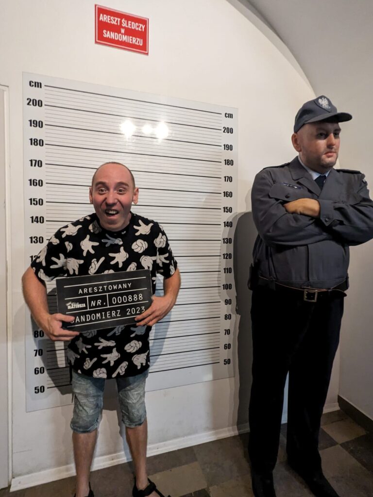 Pomieszczenie z czerwoną tabliczką "areszt śledczy w Sandomierzu". Na ścianie plansza z miarami wysokości. Przed nią stoi chłopak z tabliczką aresztowany, a obok figura woskowa policjanta. 