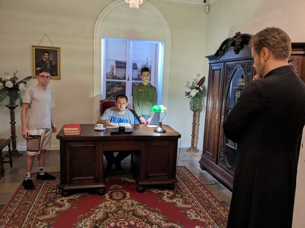 Zdjęcie w pomieszczeniu wyglądającym jak gabinet. Jeden chłopak siedzi za biurkiem. Po obu jego stronach stoją chłopcy. Przed nimi figura woskowa zamyślonego Ojca Mateusza. 