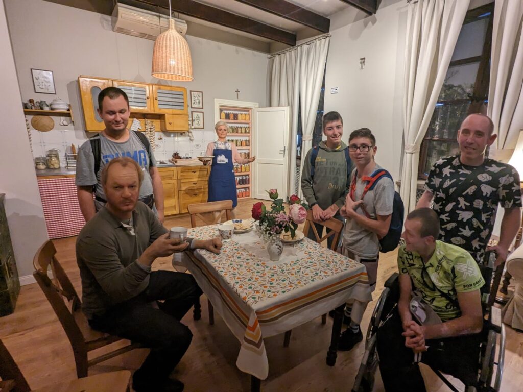 Grupa osób wokół stołu w pomieszczeniu zaaranżowanym na serialową kuchnię. Przy stole siedzi figura jednej z męskich z postaci, a w drzwiach figura gospodyni niosącej posiłek, 