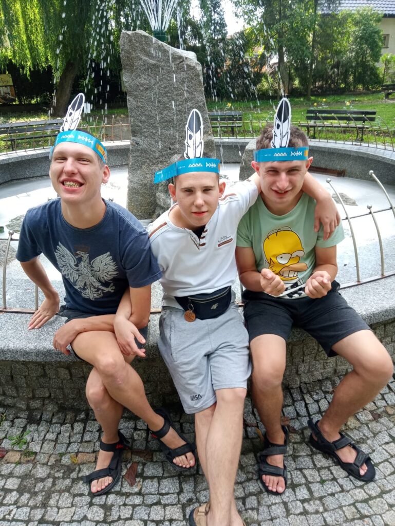Chłopcy z opaskami z piórami na głowie siedzą  przy fontannie 