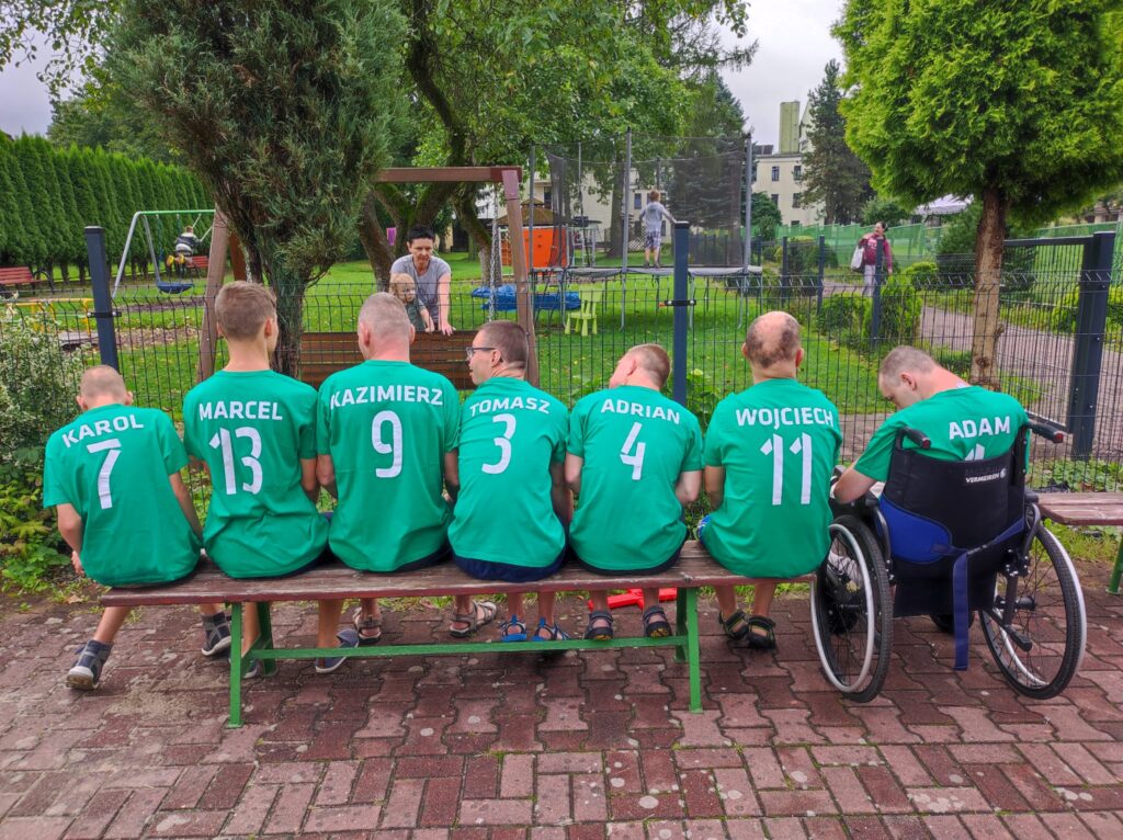 Grupa chłopaków siedząca na ławce. Zdjęcie zrobione jest od tyłu. Każdy z nich ma zieloną koszulkę ze swoim imieniem i liczbą 