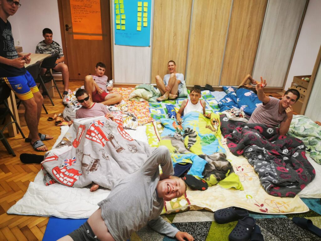 grupa chłopaków w piżamach leżących na prowizorycznych posłaniach rozłożonych na podłodze. 
