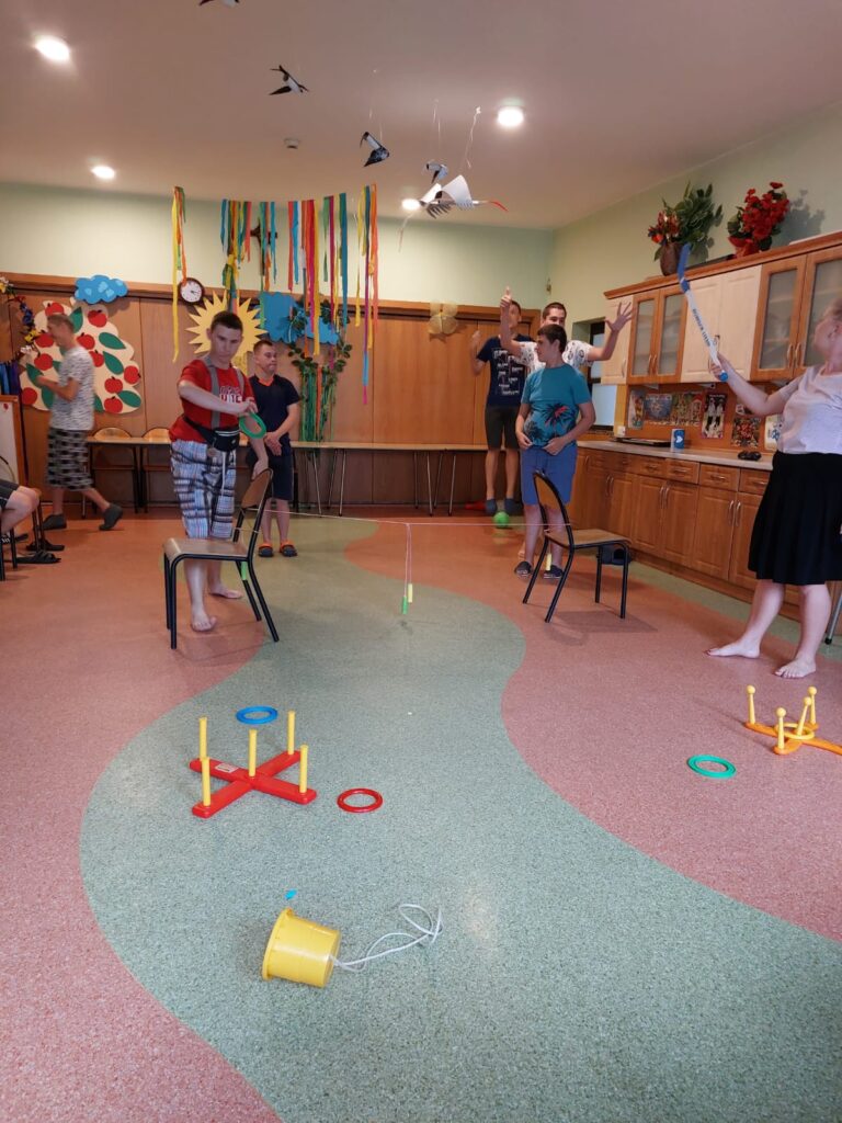Grupa osób podczas ćwiczeń w pomieszczeniu. Chłopcy rzucają okręgami do stojaków. 