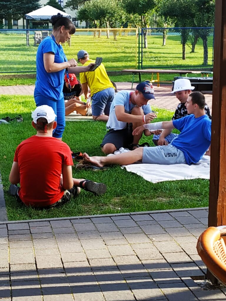 Chłopcy siedzą na kocach na trawie. Jeden bandażuje drugiemu rękę. 