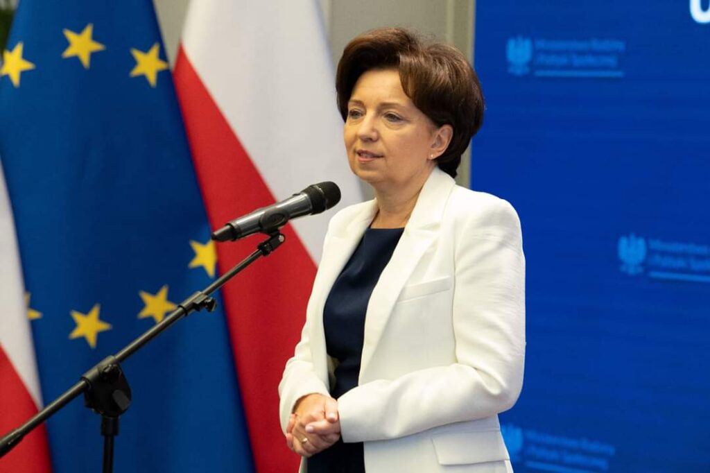 Kobieta przemawiająca do mikrofonu. Za nią stoją flagi Polski oraz Frala Uni Europejskiej. 