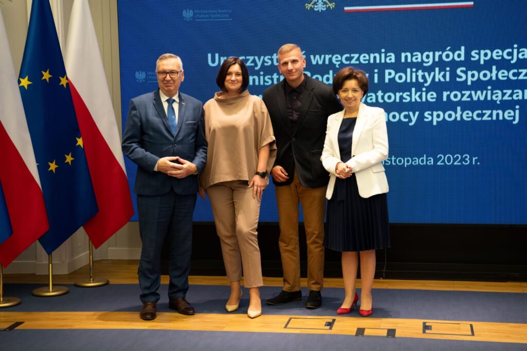 Dwie Kobiety i dwoje mężczyzn pozujących do zdjęcia tle ścianki z logo MRiPs i napisem "uroczystość wręczenia nagród specjalnych Ministra Rodziny i Polityki Społecznej za wybitne, nowatorskie rozwiązania w zakresie pomocy społecznej. Obok stoją Flagi Polski i Unii Europejskiej., 