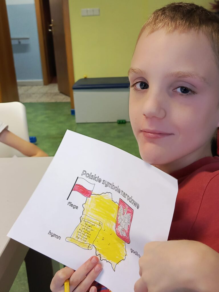 Chłopiec prezentujący swoją pracę plastyczną -kartę pracy z symbolami narodowymi. 