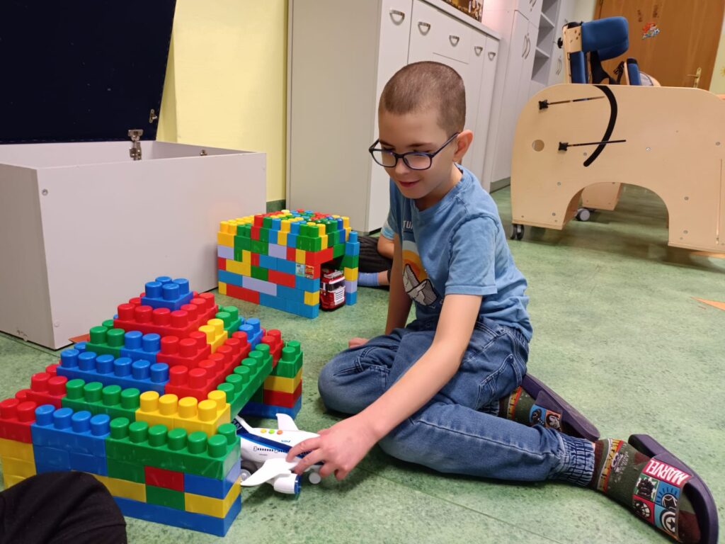 Chłopiec siedzący na podłodze. Obok niego budowle z kloców. Chłopiec wkłada do wybudowanego z klocków grażu zabawkowy samolot. 