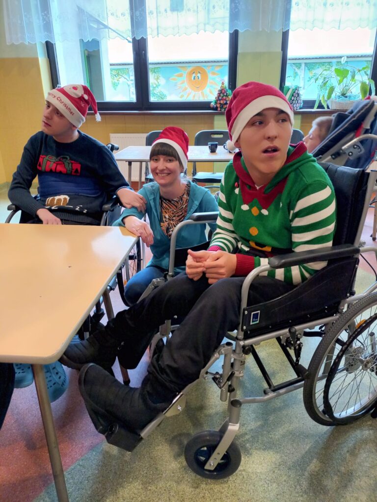 kobieta kuca pomiędzy dwoma chłopakami siedzącymi na wózkach inwalidzkich. Wszyscy mają na głowach mikołajkowe czapki. 