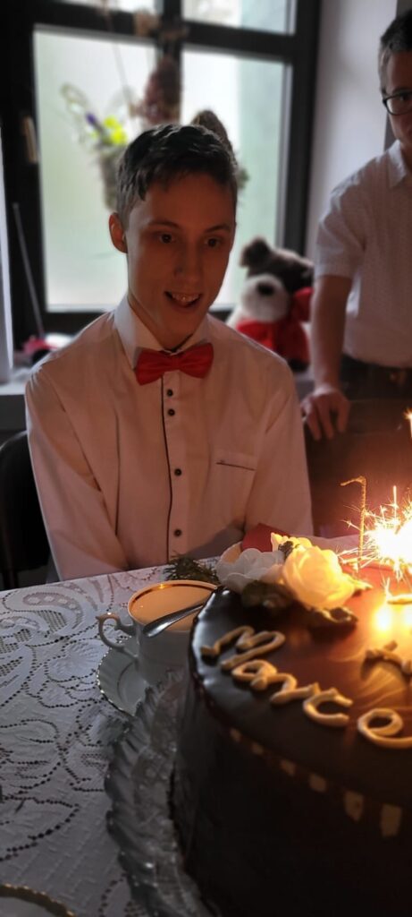 Uśmiechnięty chłopiec za stołem. Przed nim stoi tort z zapalonymi racami. 