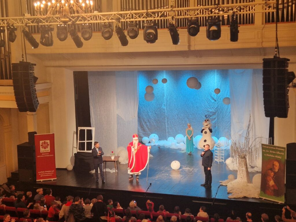 Scena z zimową aranżacją. Na scenie stoi dwoje mężczyzn w garniturach oraz aktorzy w strojach Mikołaja oraz postaci z bajki Kraina Lodu - Elsy i bałwana Olafa. 