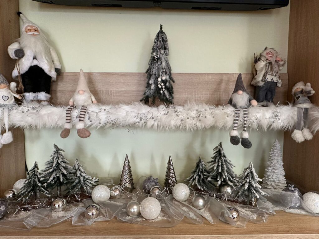 Dekoracja świąteczna: na dwóch blatach szafek (jedna nad drugą) rozłożony jest biały tiul, na którym ułożone są malutkie choineczki i złote oraz srebrne kule. Po bokach górnej szafki posadzono dwa małe, materiałowe skrzaty. 